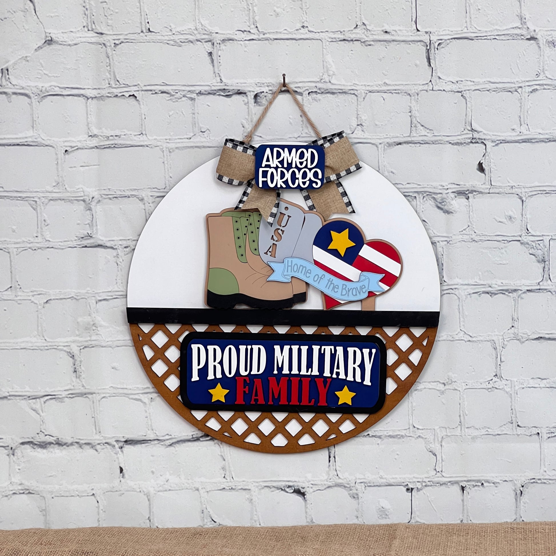 Against a white brick background, Janet's Craft Corner presents an interchangeable DIY kit: a round door hanger featuring a military boot, USA dog tag, and patriotic motifs. It reads "Armed Forces," "Proud Military Family," and "Home of the Brave" on an American-themed heart.