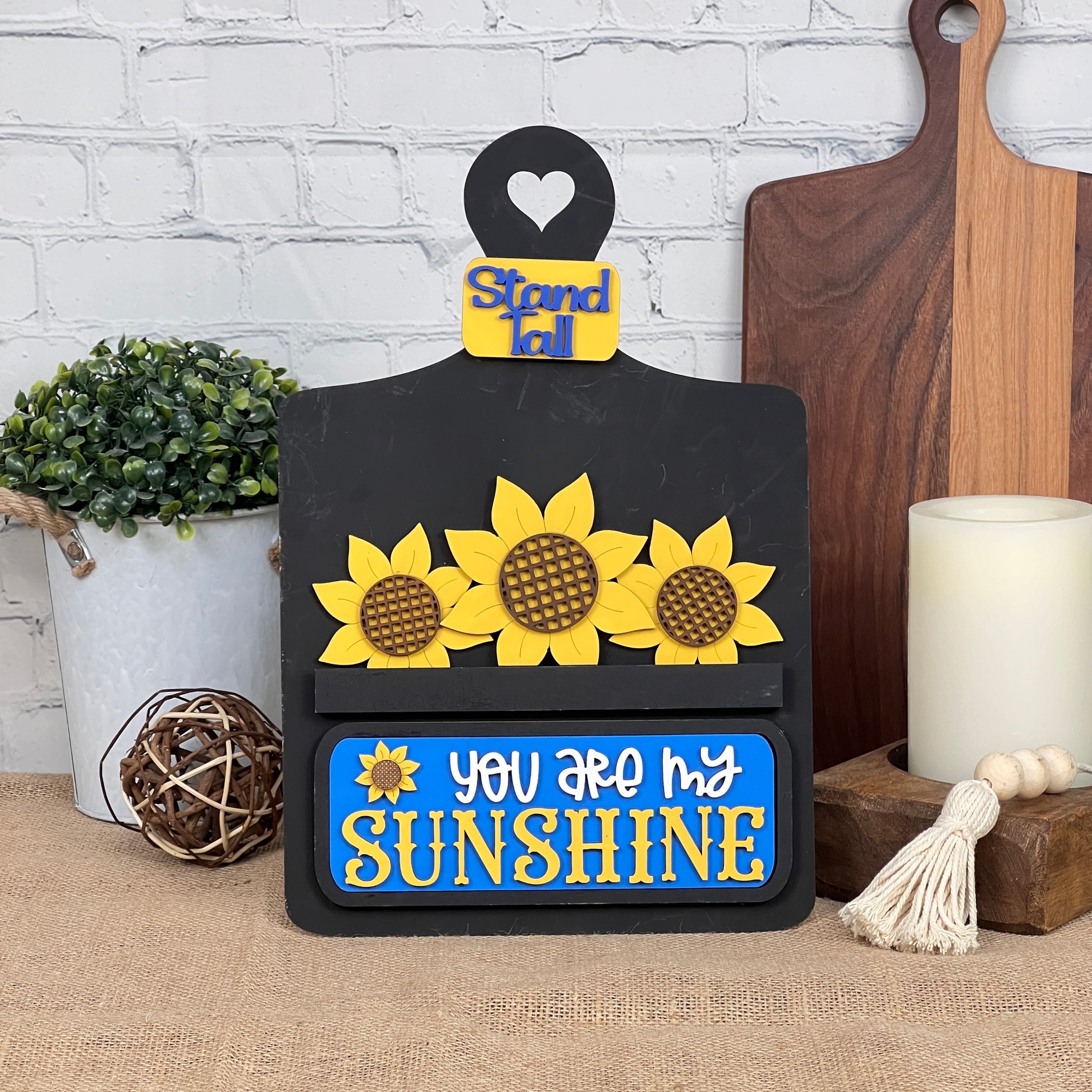 Decorate with Janet's Craft Corner's Breadboard Interchangeable Base featuring a rustic insert; perfect for home décor. Comes with a set of three sunflower embellishments and bold "You are my sunshine" text alongside "Stand tall" on a small heart. Ideal for pairing with candles, plants, and other items.