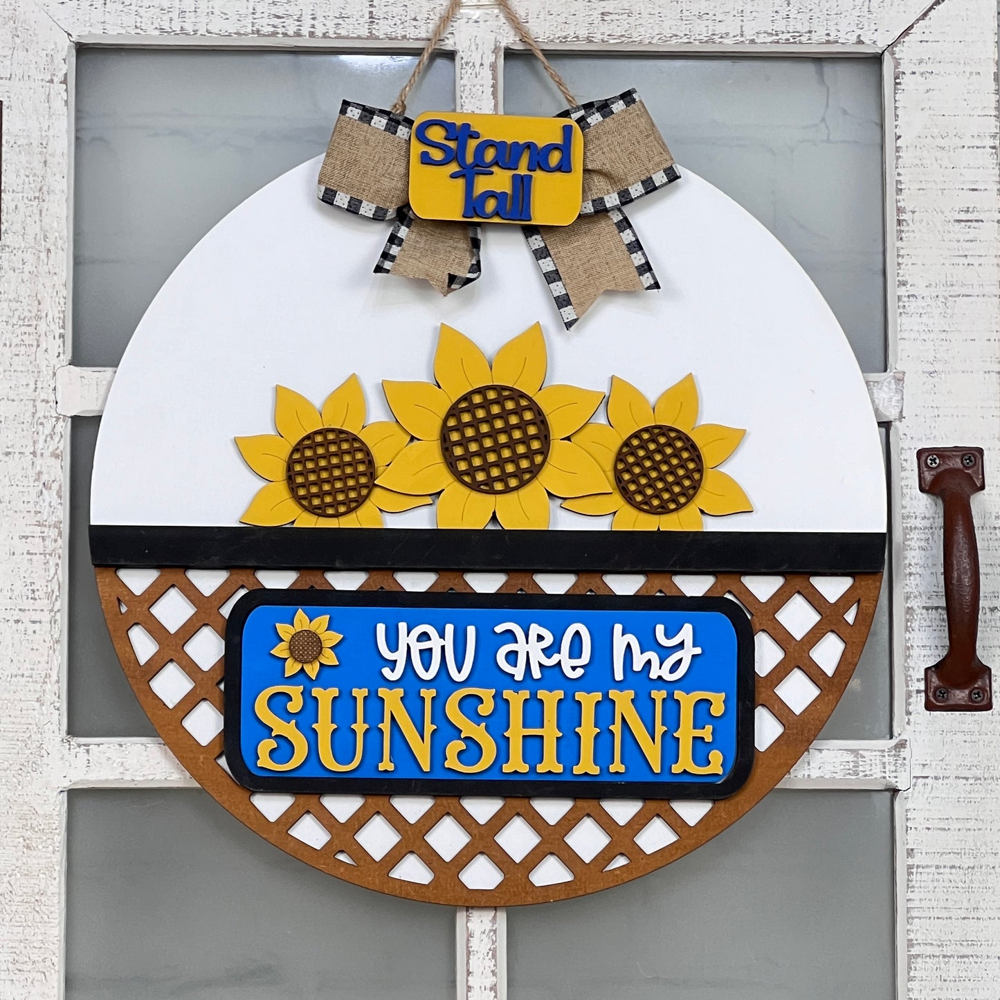 Janet's Craft Corner offers a DIY door hanger kit featuring a round base and an insert. It displays three sunflowers and the phrase "You Are My Sunshine" in yellow and white on blue. An upper sign reads "Stand Tall," accompanied by a burlap bow, making it perfect for decorative doors.