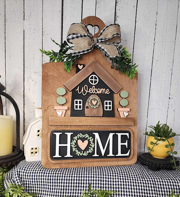 Janet's Craft Corner offers the Breadboard Interchangeable Base with Insert, a charming DIY home decor kit. Featuring a small house with "Welcome," it includes interchangeable accessories like a checkered bow and greenery, while the bold "HOME" rests on a gingham cloth backdrop.