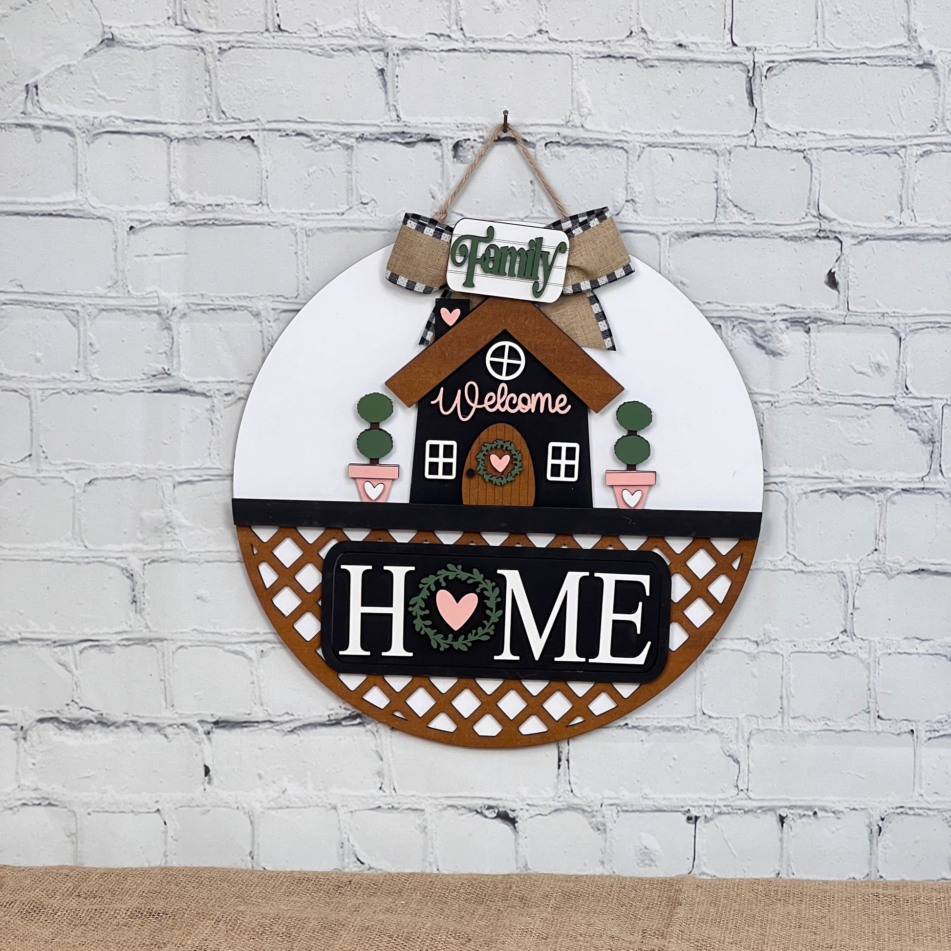 The Janet's Craft Corner Door Hanger Interchangeable Base with Insert is a circular wall decor featuring a house, "Family," and "Welcome" above, and "Home" in large letters with heart motifs below. Perfect as an interchangeable door hanger on your white brick wall.