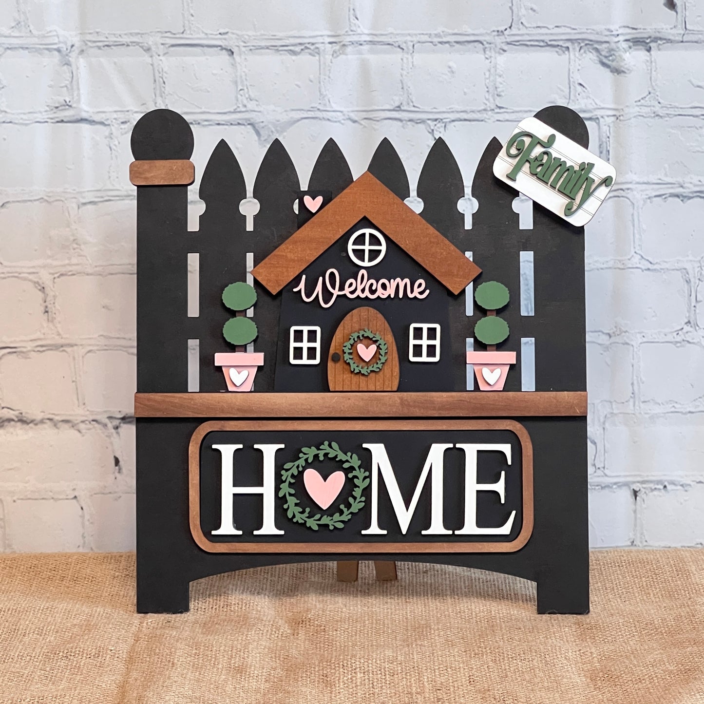 Janet's Craft Corner's Garden Fence with Insert is a DIY decor kit featuring a picket fence and house design with "Welcome" above a house with plants. "HOME" is written with a wreath as the "O," and it includes interchangeable inserts for customization, perfect against a brick wall backdrop.