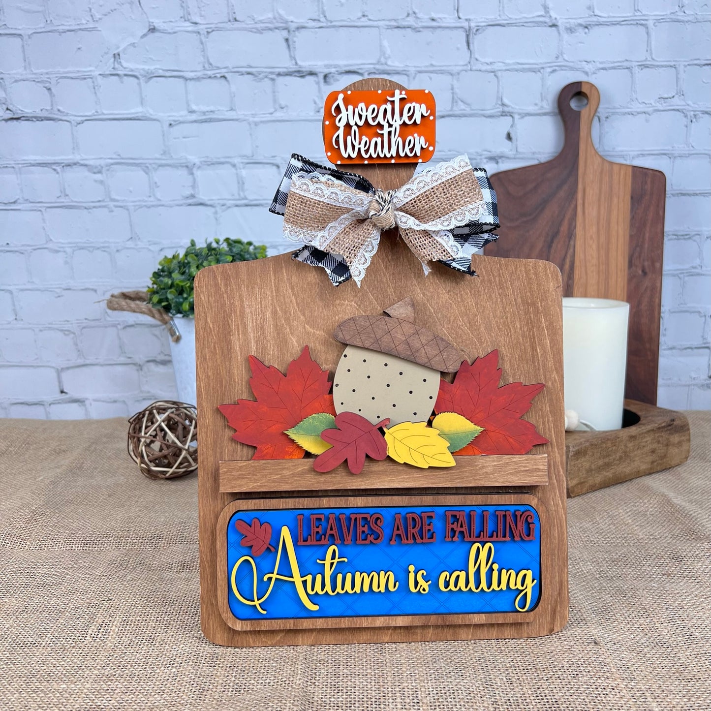 A wooden decor piece reading "Leaves are Falling, Autumn is Calling," featuring colorful leaves, an acorn, and a lace-plaid ribbon bow. Ideal for decor lovers who enjoy interchangeable items. Seen with a candle and "Sweater Weather" sign. Product: Breadboard Interchangeable Base from Janet's Craft Corner.