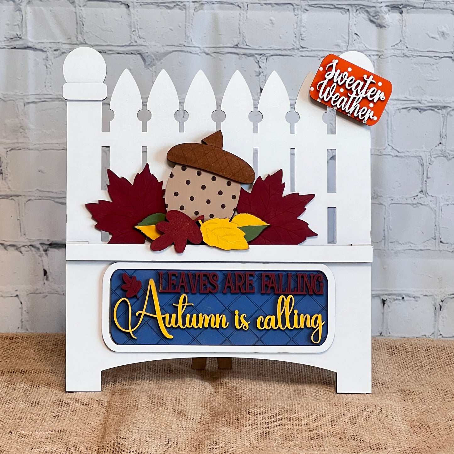 Janet's Craft Corner offers a DIY kit with a white picket fence design, featuring colorful leaves and a large acorn insert. Text reads "Leaves are Falling, Autumn is Calling" with "Sweater Weather" above, perfect for home decor against a charming brick background.