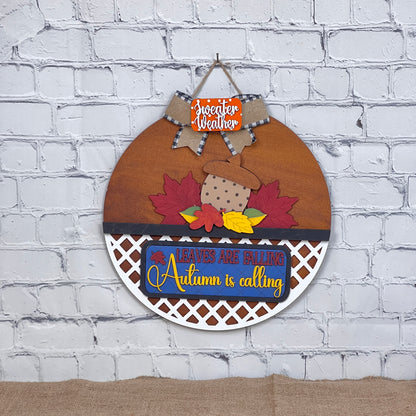 Janet's Craft Corner offers a DIY kit featuring a round wooden fall wall decoration with an acorn and leaf design. It includes an orange "Sweater Weather" sign and a blue "Leaves are Falling, Autumn is Calling" sign, ideal for interchangeable home décor on a white brick background.