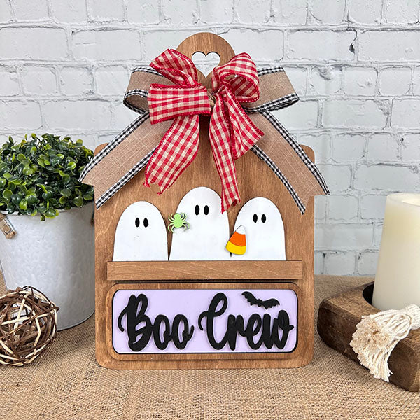 Janet's Craft Corner offers a Breadboard Interchangeable Base with Insert. This DIY kit features a wooden Halloween decor set with three ghost figures and a "Boo Crew" sign, completed by a red and black plaid ribbon bow, interchangeable accessories, potted plant, and candle against a white brick background.