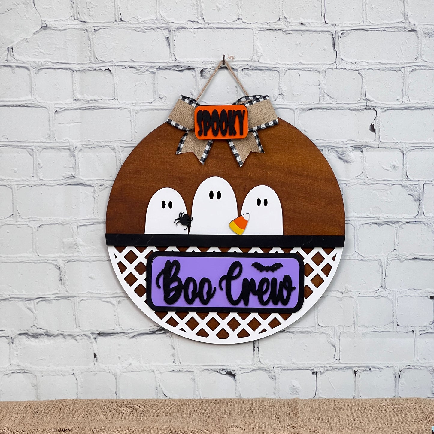 Janet's Craft Corner offers a DIY interchangeable home décor kit featuring a "Boo Crew" round wooden sign with three cartoon ghosts, purple lettering, and "Spooky" in orange on burlap. Includes charming details like tiny black eyes and a spider against a brick wall, perfect for door decor enthusiasts.