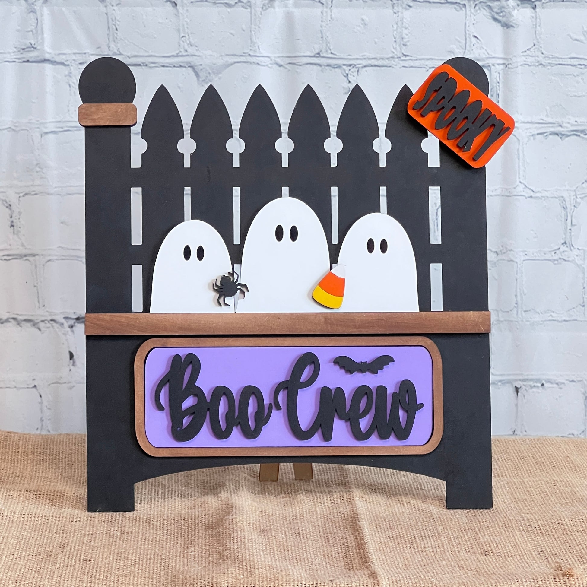 The Garden Fence with Insert from Janet's Craft Corner features three white ghosts behind a black picket fence with a "Boo Crew" plaque, spider and candy corn detail, and an orange "Spooky" sign. Perfect for DIY home decor against a white brick wall.