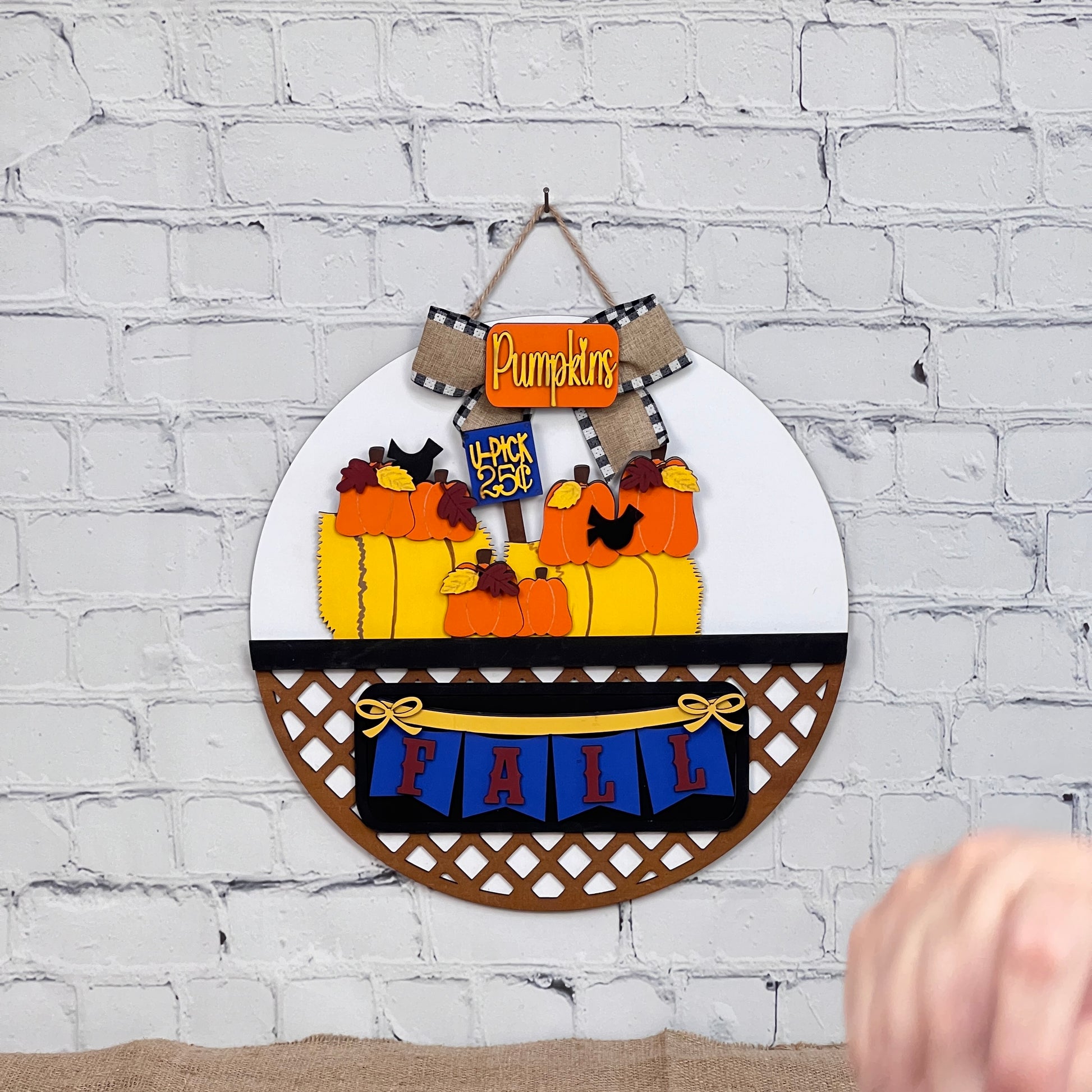 A round wall piece from Janet's Craft Corner decorates a white brick wall, enhancing your home with pumpkins and a "3 for $10" sign. A "FALL" banner adorns the base, and it features an interchangeable door hanger option for versatility. A hand is partially visible at the bottom.