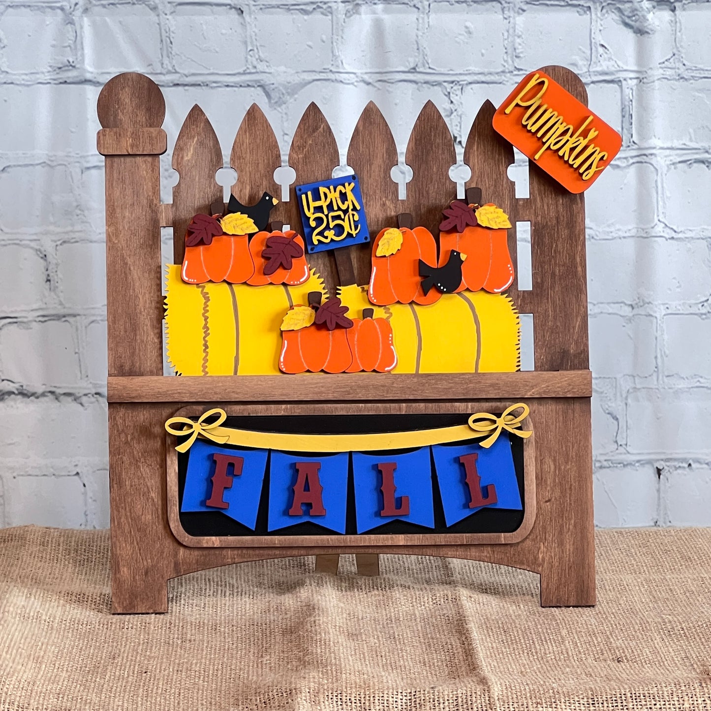 Janet's Craft Corner presents a delightful DIY home decor kit featuring a Garden Fence with an insert. It includes a wooden picket fence decorated with paper pumpkins, a "Pumpkins" sign, and "4 Pack 25¢" label, completed by a blue and red "FALL" banner set against a rustic white brick wall.