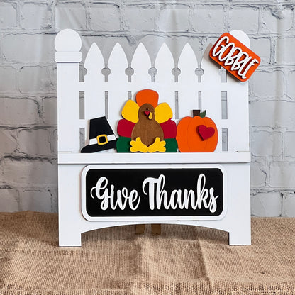 Janet's Craft Corner presents the Garden Fence with Insert, a DIY home decor kit featuring a white picket fence with a felt turkey, pilgrim hat, and pumpkin. "Gobble" adorns a top sign and "Give Thanks" graces the bottom plaque, ideal for crafting against any brick wall setting.