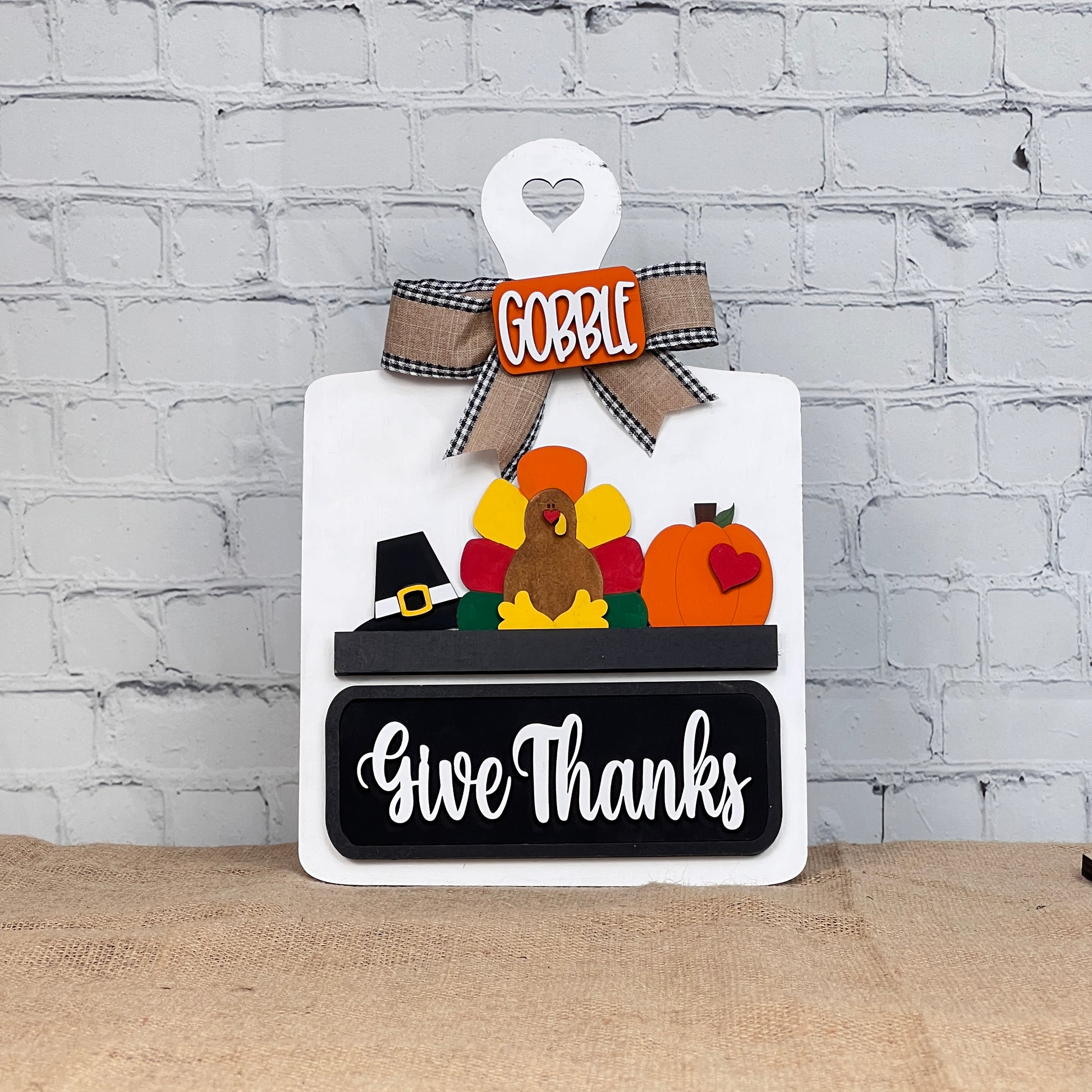 The "Breadboard Interchangeable Base with Insert" from Janet's Craft Corner features a turkey with colorful feathers, a pilgrim hat, and the words "Gobble" and "Give Thanks." This delightful DIY home decor piece rests against a white brick wall on burlap.