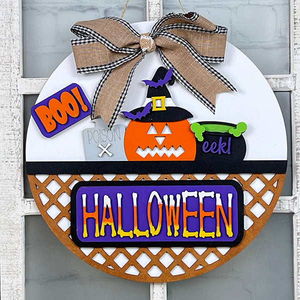 Janet's Craft Corner offers a DIY kit with a round Halloween decoration: a witch-hat pumpkin, "poison" potion bottle, green cauldron, "BOO!" and "eek!" signs, topped with a plaid bow. Perfect as an interchangeable door hanger for festive home decor!.