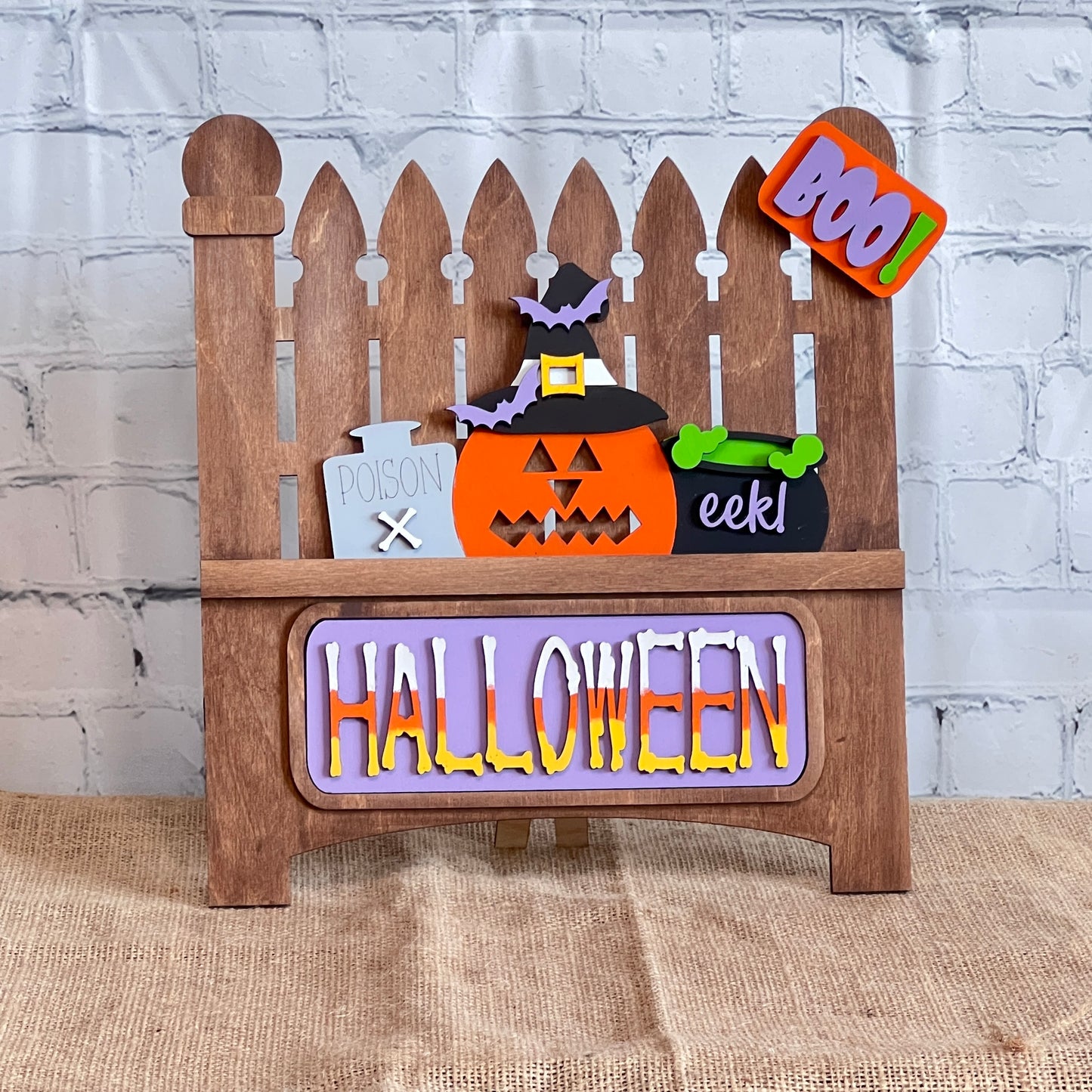 The Garden Fence with Insert from Janet's Craft Corner is a charming wooden Halloween decor featuring a picket fence design. It includes interchangeable elements like a pumpkin in a witch hat, "POISON" sign, green "eek!" cauldron, and colorful "BOO!" and "HALLOWEEN" signs, perfect for DIY lovers.