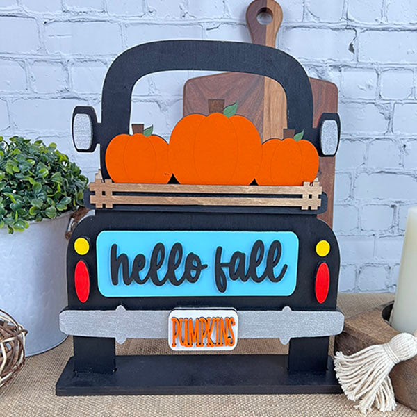 Janet's Craft Corner's Antique Truck Base with Insert, a DIY interchangeable home decor kit, features a charming decorative sign. Shaped like an antique truck carrying four pumpkins labeled "hello fall" and includes a "PUMPKINS" plate, perfect for display against a white brick wall with plants and candles nearby.