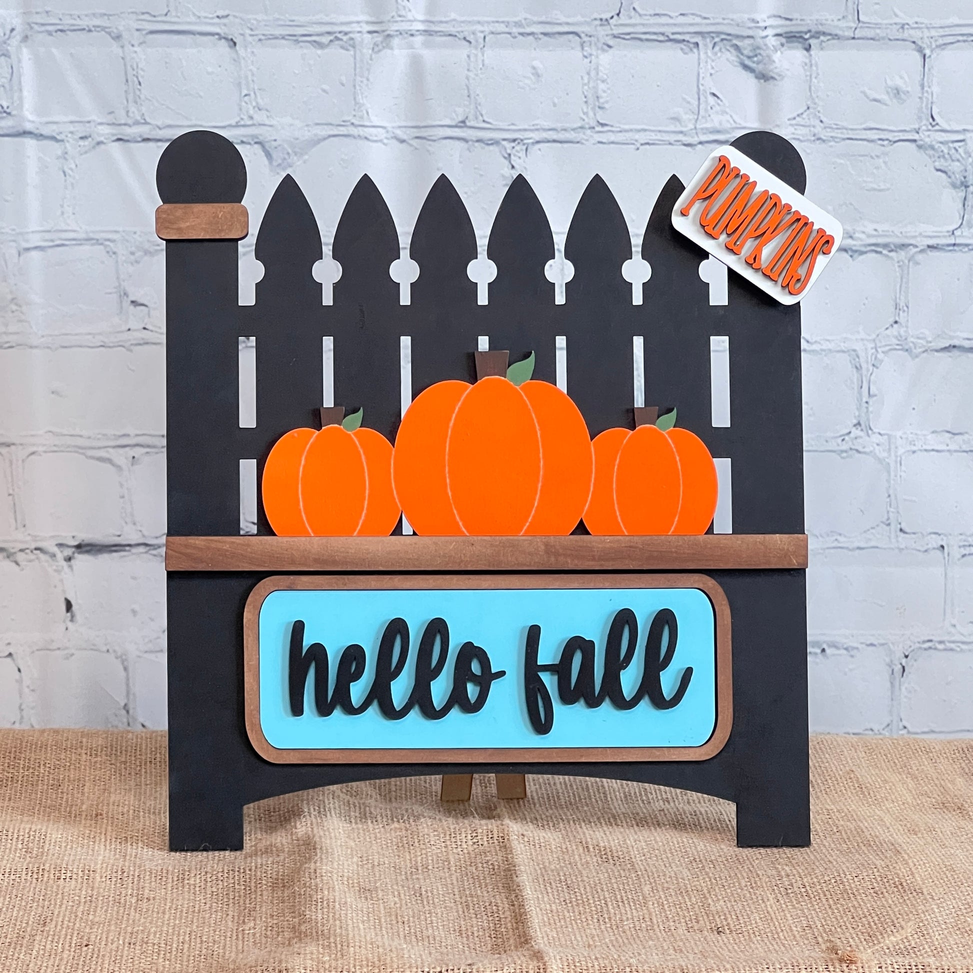 A decorative sign by Janet's Craft Corner showcases three orange pumpkins from the Garden Fence with Insert craft kit. Against a white brick wall, it displays "hello fall" in blue and "Pumpkins" on a small sign above, with an interchangeable Picket Fence silhouette as the backdrop.