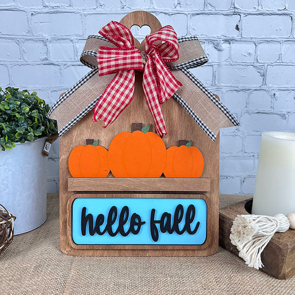 The Breadboard Interchangeable Base by Janet's Craft Corner is a DIY kit featuring a charming breadboard with three orange pumpkins and "hello fall" sign. It’s topped with a red and white checkered bow, set against a light brick wall, with a plant and candle in the background.