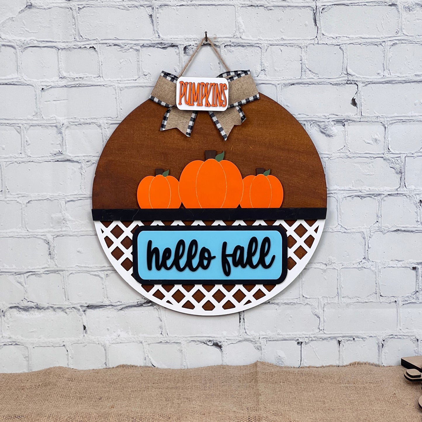 The Door Hanger Interchangeable Base with Insert from Janet's Craft Corner features a round wooden design with "PUMPKINS," a bow, vibrant pumpkins on lattice, and a blue "hello fall" backdrop. This DIY home décor kit is perfect for warmly greeting guests on your white brick wall.