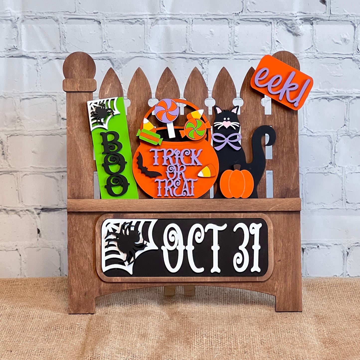 Janet's Craft Corner offers a Garden Fence with Insert, featuring a wooden Halloween-themed design adorned with "Boo," "Trick or Treat" signs, a black cat, pumpkin, lollipops, and spider web against a white brick wall—perfect for festive home decor.