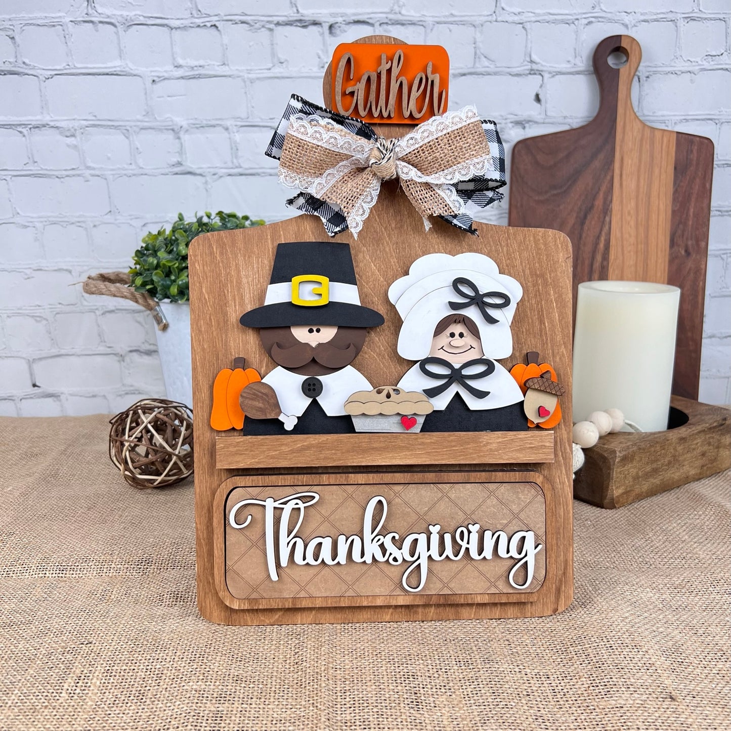 The Breadboard Interchangeable Base with Insert by Janet's Craft Corner is a DIY kit featuring cartoon Pilgrims, pumpkins, a pie, and "Thanksgiving" in white script atop a wooden panel. It includes interchangeable accessories like candles and cutting boards for rustic home decor enhancement.