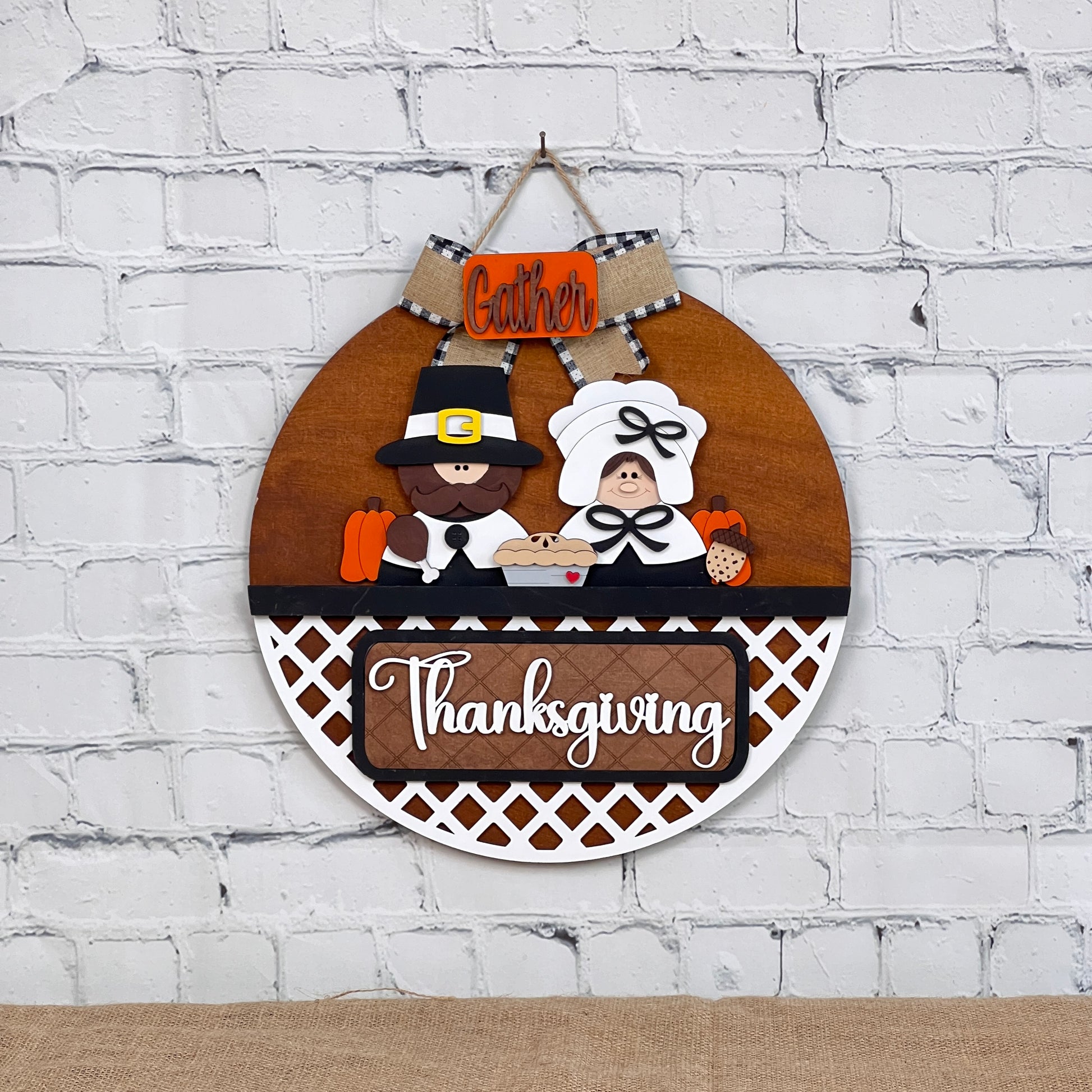 Janet's Craft Corner offers a round wooden Thanksgiving sign featuring "Gather” and "Thanksgiving" text. A pilgrim couple holds a pie and a pumpkin against a white brick background with a lattice pattern below. Perfect for DIY craft enthusiasts, this interchangeable door hanger includes one base and insert.