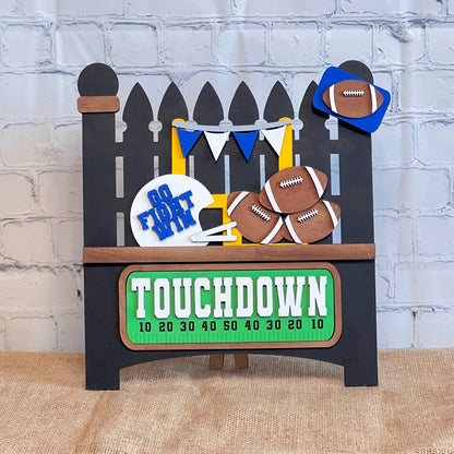 Janet's Craft Corner offers a Garden Fence with Insert DIY kit featuring a football-themed display. It includes a picket fence with pennants, footballs, and a "Go Fight Win" sign above a burlap surface. A green scoreboard-style sign reads "TOUCHDOWN," all set against a rustic brick wall backdrop.