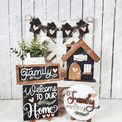 Janet's Craft Corner offers the Family Home Decor Set, a DIY kit featuring personalized wooden signs reading “Family,” “Welcome to Our Home,” and “Family is Everything.” It includes a house-shaped plaque, green plant, and garland with small houses and hearts against a white backdrop.