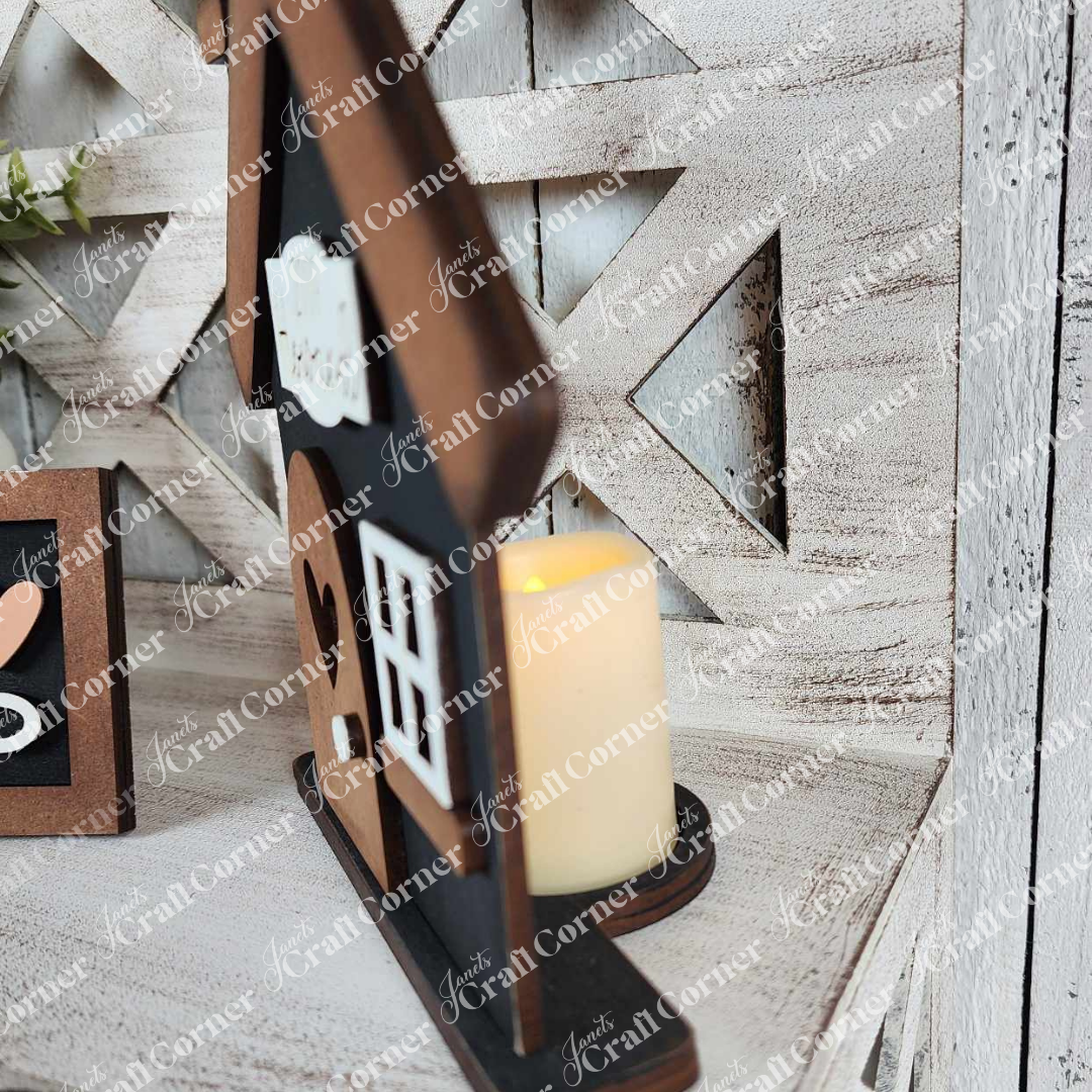 A close-up showcases Janet's Craft Corner's Family Home Decor Set, a wooden house-shaped piece with a cutout window. A warm LED candle glows behind it against a rustic crisscross wooden plank background, enhanced by a text watermark adding charm.