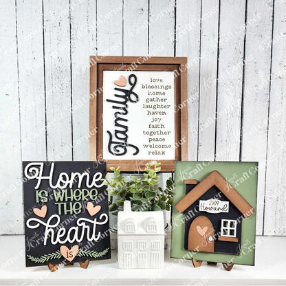 The House Interchangeable Farmhouse Sign by Janet's Craft Corner is displayed against a white wooden backdrop with words like "family" and "home," complemented by a ceramic house figurine, perfect for a DIY craft kit project.