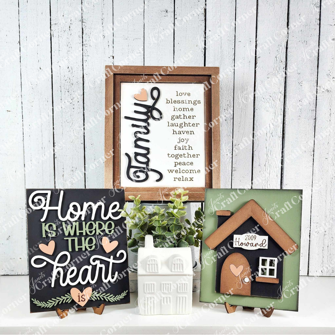 Displayed against a white plank wall, signs from Janet's Craft Corner exude rustic charm. One reads "Family," featuring words like "love" and "blessings," while another says, "Home is where the heart is." A ceramic house and a farmhouse sign complete this cozy Home Heart Interchangeable kit.