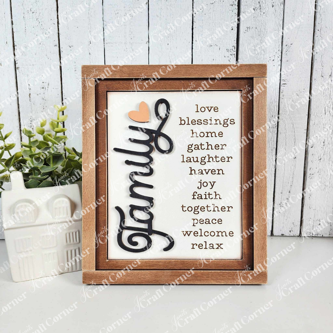 The Family Words Farmhouse Sign by Janet's Craft Corner features a wooden frame with "Family" prominently in cursive, surrounded by words like love, blessings, laughter, and peace. It pairs beautifully with a white ceramic house and green plant on whitewashed wood panels.