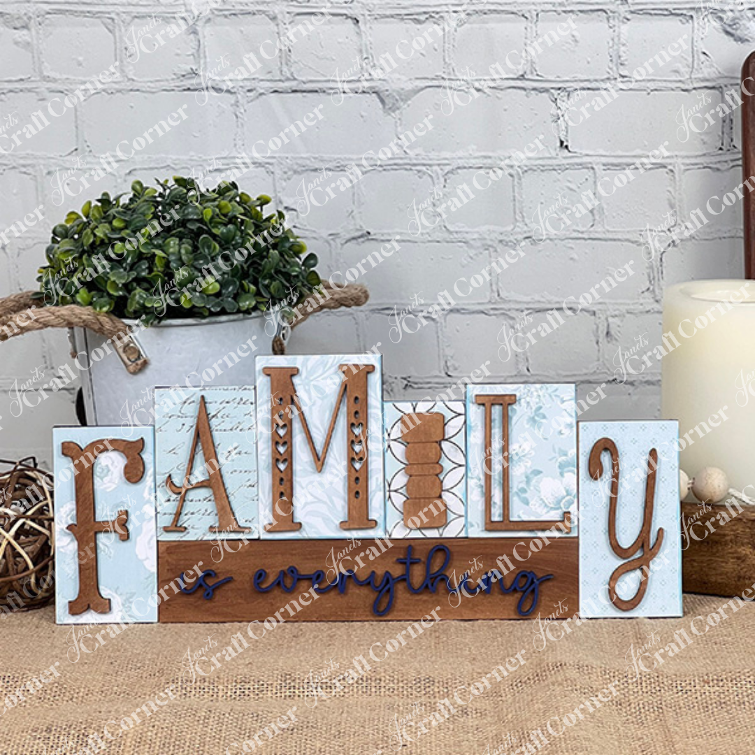 The "Family is Everything" Word Block DIY Home Decor Craft Kit from Janet's Craft Corner features decorative wooden blocks with floral patterns and rustic accents, perfect for enhancing any home decor against a burlap-covered surface and white brick wall, complete with a potted plant and rope.