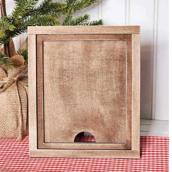 A DIY home decor craft kit featuring the Farmhouse Frame for Interchangeable Signs from Janet's Craft Corner stands upright on a checkered red and white cloth. In the background, there's a potted evergreen tree wrapped in burlap, adding a touch of farmhouse decor to the scene.