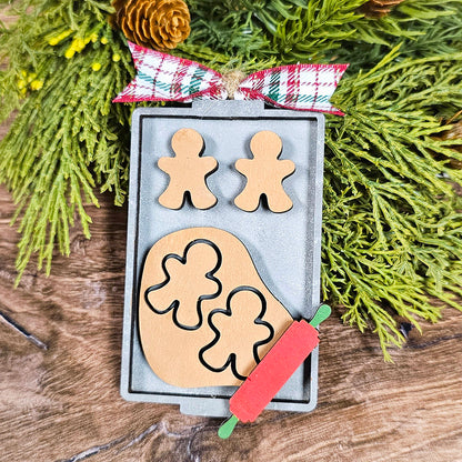 A festive scene with gingerbread men and clover shapes on a baking tray, surrounded by greenery and a plaid ribbon. A small red rolling pin is placed nearby on a wooden surface, reminiscent of crafting your own Janet's Craft Corner Baking Spirits Bright Ornaments - Individual Painted DIY kit.