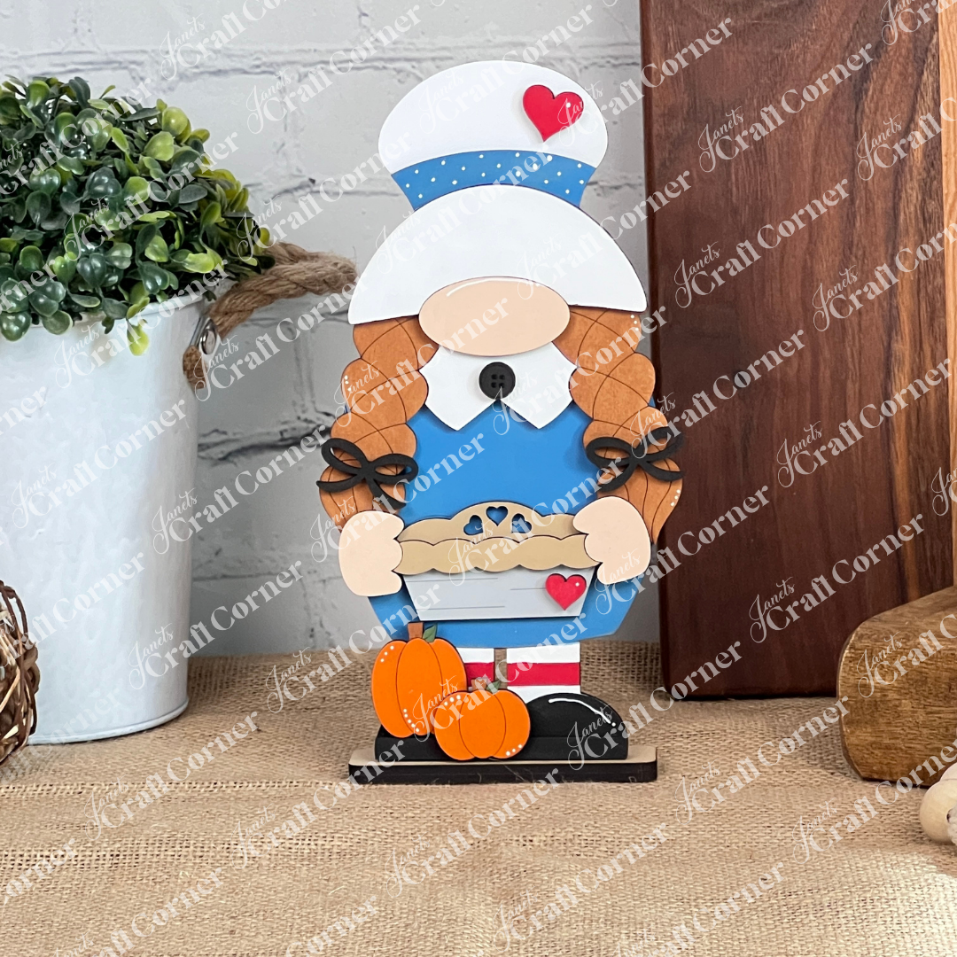 The "Girl Pilgrim Gnome Shelf Sitter" from Janet's Craft Corner features a charming wooden figure of a girl in a blue dress and white chef hat, holding a pie adorned with a heart cutout. Her braided hair complements the red heart detail on her hat, and she stands on a base decorated with small pumpkins, making it an endearing addition to personalized home décor. A nearby plant rests on a burlap-covered surface for added rustic charm.