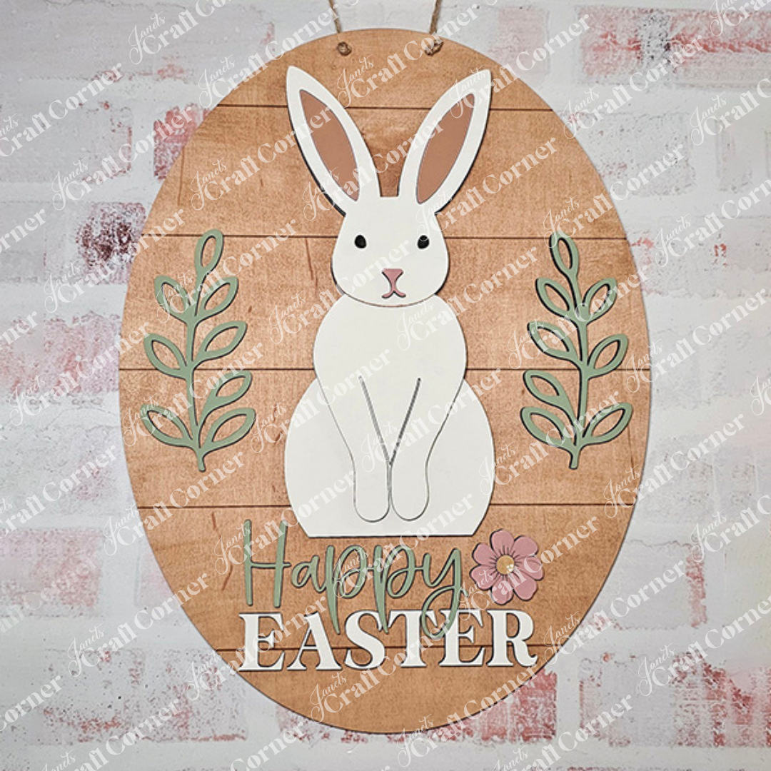 The "Happy Easter Door Hanger" from Janet's Craft Corner is a wooden decoration with a charming white bunny with long ears nestled among green leaves. The phrase "Happy Easter" appears elegantly in white beneath, accented by a small pink flower.