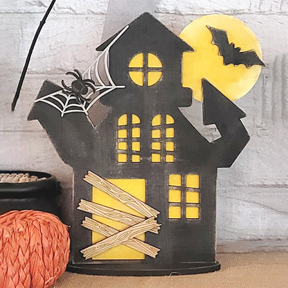 From Janet's Craft Corner, this Haunted Mansion Shelf Sitter offers a Halloween vibe with boarded windows, a spider on its web, and a bat soaring beneath the full moon. An orange pumpkin adds to the scene, complemented by a mysterious black item to complete the spooky atmosphere.