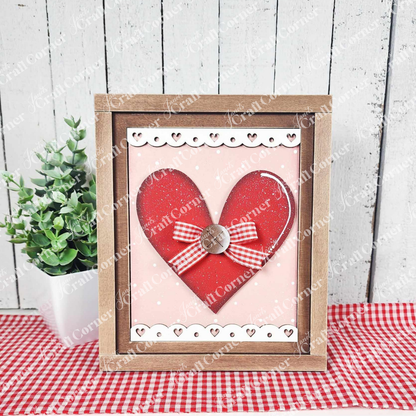 The Heart Interchangeable Sign by Janet's Craft Corner is a framed DIY craft kit featuring a large red heart with a button and bow. The decorative border complements the wooden background, and it's beautifully accented with a small green plant and red-checked cloth for charming home decor.