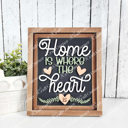 Janet's Craft Corner's Home Heart Interchangeable Sign reads "Home is where the heart is" in stylized white and green text with small hearts. Perfect for home décor, it’s propped beside a plant and ceramic house on a light wooden background, adding charm to any space.