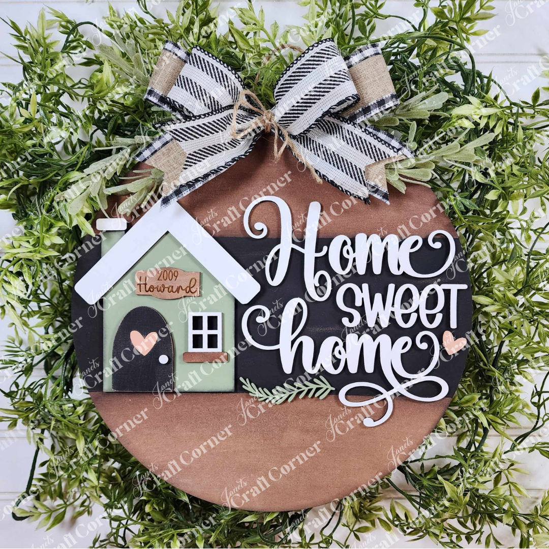 The Home Sweet Home Door Round by Janet's Craft Corner features a hand-painted circular wooden sign with "Home Sweet Home" in white cursive. A charming house with a green mat and bow sits atop, encircled by greenery for a wreath effect, making it an ideal DIY craft kit for home décor lovers.