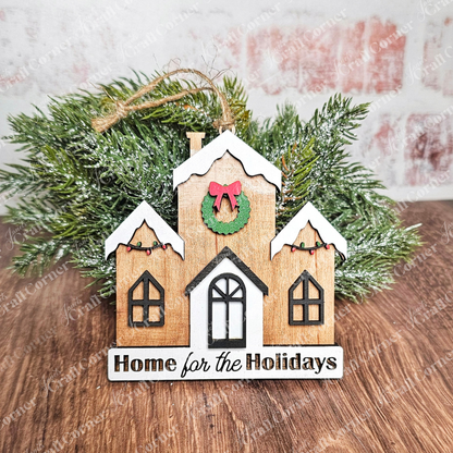 Introducing the "Holiday Village Ornament Set - Early Access" from Janet's Craft Corner: a delightful, hand-painted decor piece shaped like a snow-covered house adorned with a wreath, featuring a "Home for the Holidays" sign. It is set against green, frosted pine branches on a brick surface, making it an exquisite addition to any Holiday Village Ornament Set.