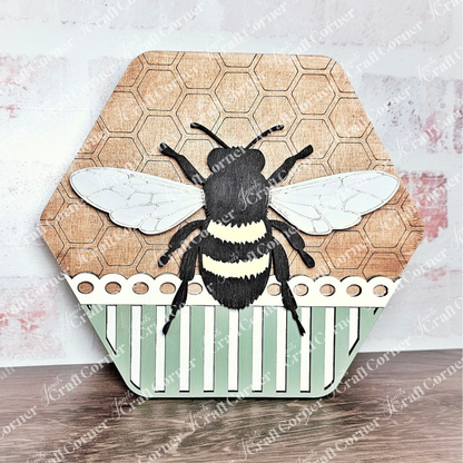 The Honeycomb Shaped Bee Sign from Janet's Craft Corner is a wooden hexagonal plaque ideal for DIY projects, featuring a painted bee with intricate wings. The upper half has a honeycomb pattern, while the lower half showcases vertical green and white stripes, perfect for any Bee Sign DIY craft kit.