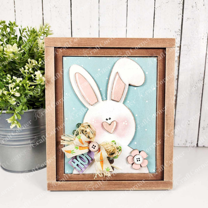 Janet's Craft Corner presents the "Hop Bunny Farmhouse Sign," a wood-framed art piece of a white bunny with pink ears and nose on a light blue background, holding a "HOP" sign. The design includes flowers with button centers, reminiscent of a charming seasonal craft kit, alongside a potted plant.