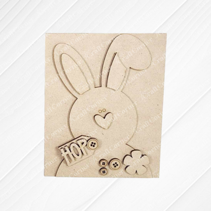 The "Hop Bunny Farmhouse Sign" by Janet's Craft Corner features a wooden bunny with a heart-shaped nose and large ears, perfect for DIY decor. It has "HOP" engraved on its side with button and flower shapes against a textured white background.