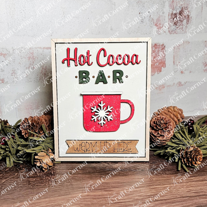 The "Hot Cocoa Bar" Interchangeable Sign by Janet's Craft Corner shows a red mug and snowflake design with pine cones and greenery, complemented by a "Warm Up Here" banner against a whitewashed brick backdrop, ideal for any DIY craft kit display.