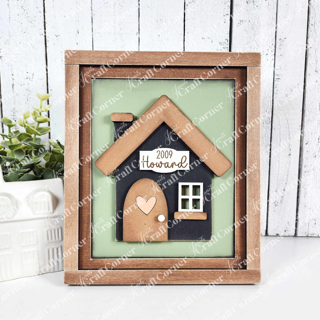 A charming DIY craft kit from Janet's Craft Corner features a farmhouse sign with a cozy house and heart door, "2009 Howard" text on a light green background. A plant and decor enhance the inviting scene in this House Interchangeable Farmhouse Sign set of 1.