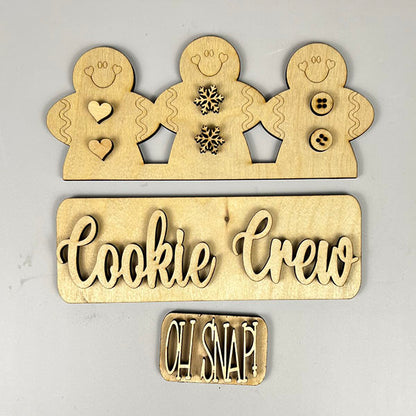 The Cookie Crew Interchangeable Insert by Janet's Craft Corner is an ideal DIY home decor kit, featuring wooden cutouts of three gingerbread men adorned with heart, snowflake, and button designs. A sign at the base displays "Cookie Crew" with a smaller "Oh Snap!" text, all crafted from light-colored wood.