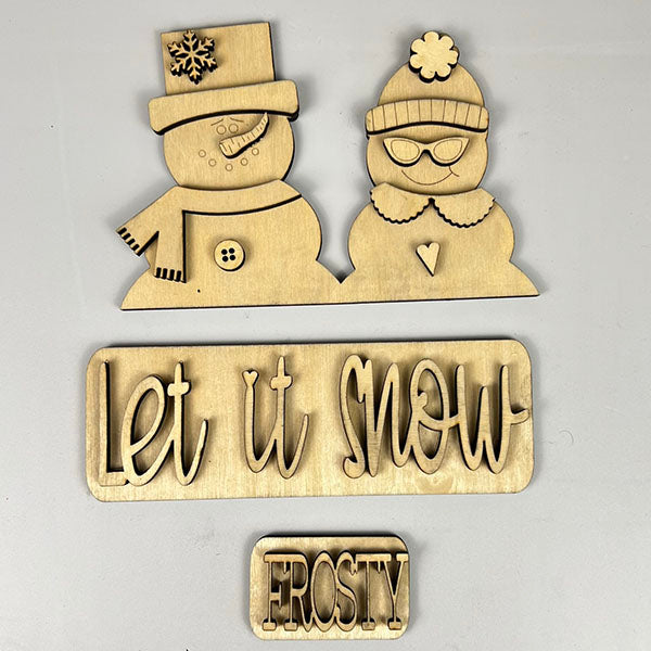 Janet's Craft Corner presents a DIY home decor kit featuring wooden cutouts of a snowman couple, with one donning a snowflake hat and the other wearing a beanie and glasses. Below them are decorative fonts displaying "Let it Snow" and "FROSTY," making up the Let It Snow Interchangeable Insert.