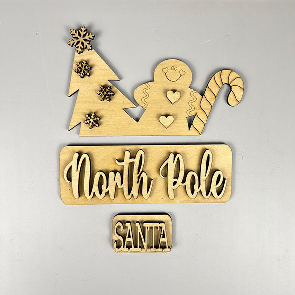 The "North Pole Interchangeable Insert" by Janet's Craft Corner is a delightful wooden cutout decoration showcasing a Christmas tree adorned with snowflakes, accompanied by a gingerbread figure and a candy cane. Below, the words "North Pole" are written, with a smaller piece labeled "Santa." This charming piece is designed as an interchangeable insert for your DIY home decor collection.