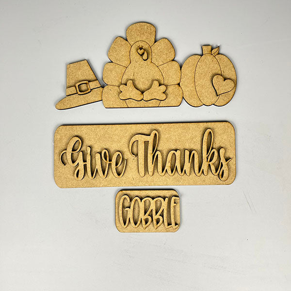 Enhance your home decor with Janet's Craft Corner's "Give Thanks Interchangeable Insert" DIY home decor kit. It features wooden cutouts showcasing a turkey, pilgrim hat, and pumpkin, along with the phrases "Give Thanks" and "Gobble," all set against a white background.